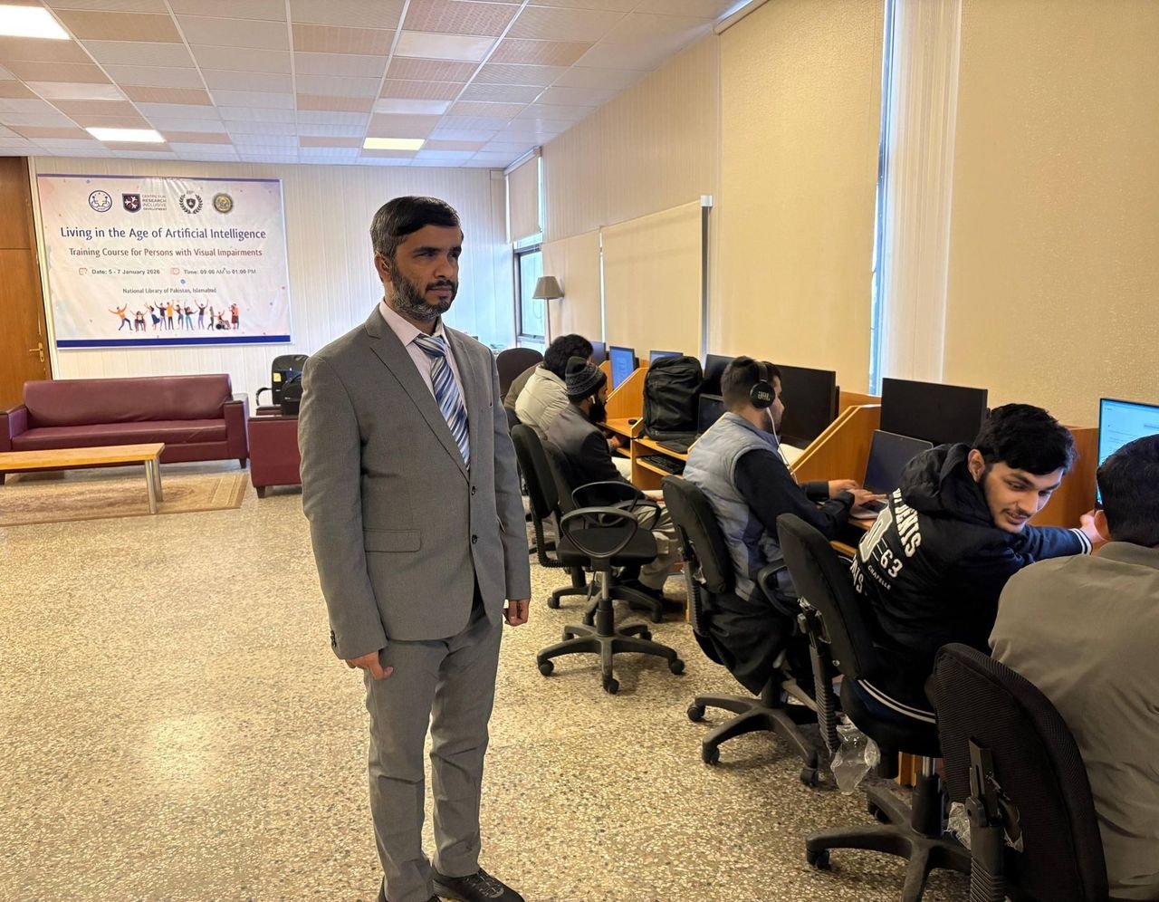 A man in a gray suit and striped tie stands in a computer lab during the “Living in the Age of Artificial Intelligence: Training Course for Persons with Visual Impairments” at the National Library of Pakistan. Behind him, several participants with headphones work at desktop computers in a row of wooden cubicles, while a banner on the back wall displays the event title, dates, and colorful silhouettes of people celebrating.