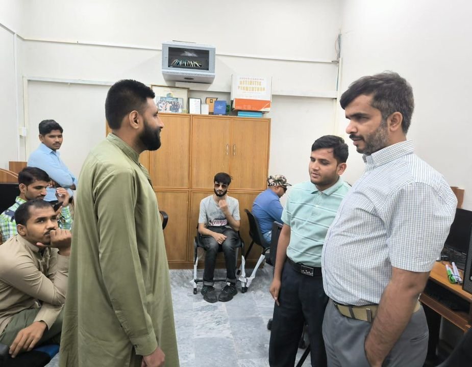 A group of men are gathered in a small computer lab at the PFFB complex during a “Training for Accessible Call Center Careers” session. Three participants stand in the center of the room in conversation, while others sit at computer workstations along the walls, listening and working. The room has light-colored walls, a wooden storage cabinet at the back with documents and boxes on top, and several desktop computers on wooden desks, giving the scene a focused, classroom-style training atmosphere