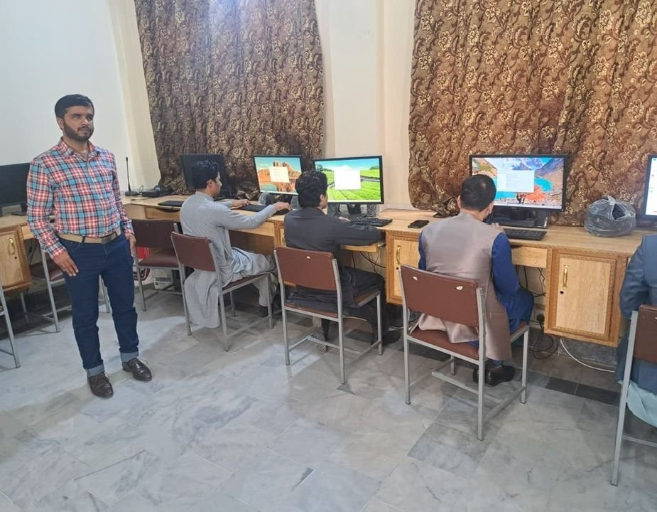 A group of men participate in a “Training of Trainers for Accessible Digital Education” session in Peshawar, seated at a row of desktop computers in a classroom-style computer lab. Four trainees with medium skin tones face the monitors and work individually, while an instructor in a red and blue checked shirt and dark jeans stands to the left, facing the camera. The room has light-colored tile flooring, wooden computer desks, and large patterned curtains covering the back wall, creating a focuse