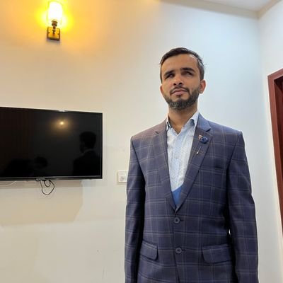 Abdullah stands indoors against a light-colored wall, wearing a blue checked suit with a light patterned shirt and a decorative lapel pin, giving him a formal, polished appearance. He has medium brown skin, short dark hair, and a trimmed beard, with his head slightly turned and a thoughtful expression on his face. To his left, a wall-mounted flat-screen TV and a glowing wall sconce are visible, and to his right there is a wooden door with raised panels.