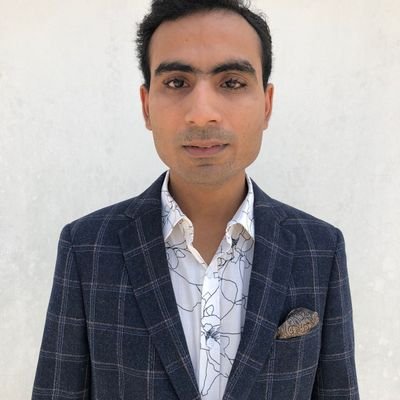Waqas, a man with medium brown skin and short, slightly tousled dark hair, faces the camera with a neutral expression against a plain light background. He is dressed in a dark checked blazer with a patterned pocket square and a white shirt featuring a fine black floral line design, giving him a smart, formal appearance.