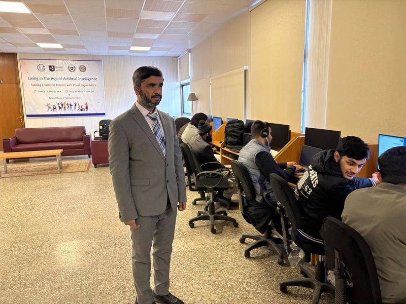 A man in a gray suit and striped tie stands in a computer lab during the “Living in the Age of Artificial Intelligence: Training Course for Persons with Visual Impairments” at the National Library of Pakistan. Behind him, several participants with headphones work at desktop computers in a row of wooden cubicles, while a banner on the back wall displays the event title, dates, and colorful silhouettes of people celebrating.