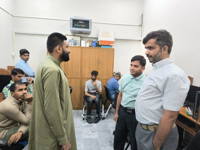 A group of men are gathered in a small computer lab at the PFFB complex during a “Training for Accessible Call Center Careers” session. Three participants stand in the center of the room in conversation, while others sit at computer workstations along the walls, listening and working. The room has light-colored walls, a wooden storage cabinet at the back with documents and boxes on top, and several desktop computers on wooden desks, giving the scene a focused, classroom-style training atmosphere