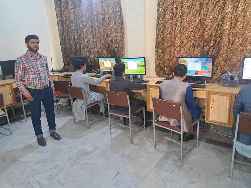 A group of men participate in a “Training of Trainers for Accessible Digital Education” session in Peshawar, seated at a row of desktop computers in a classroom-style computer lab. Four trainees with medium skin tones face the monitors and work individually, while an instructor in a red and blue checked shirt and dark jeans stands to the left, facing the camera. The room has light-colored tile flooring, wooden computer desks, and large patterned curtains covering the back wall, creating a focuse
